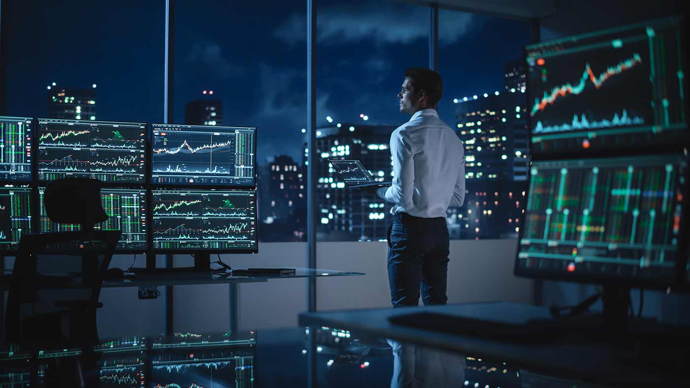 Businessman standing staring out window contemplating trades computer screens with candlestick chart patterns on desks around him