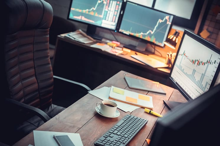 Day trader desk showing various candlestick market graphs on computer screens notebook with post-its and coffee cup on desk