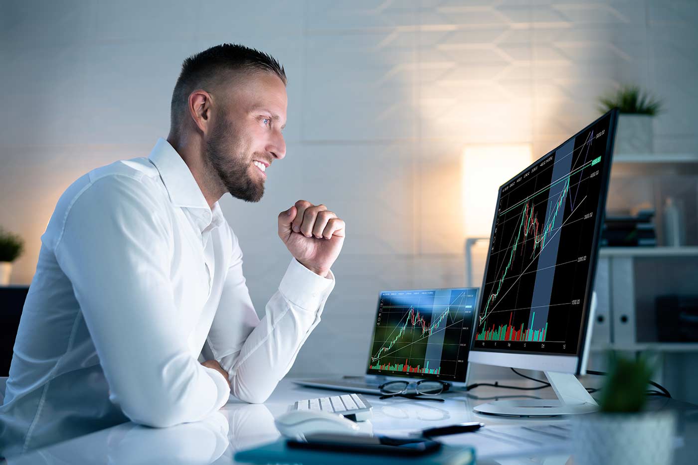 A young businessman in a white shirt sitting at a desk watching a computer and celebrating day trader win