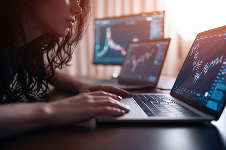 Female day trader leaning forward looking at stock charts on laptop with two computer screens in background