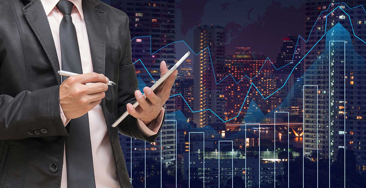 A business man in a grey suit holds a tablet with a pen, while the city background is overlaid with a trading graph.