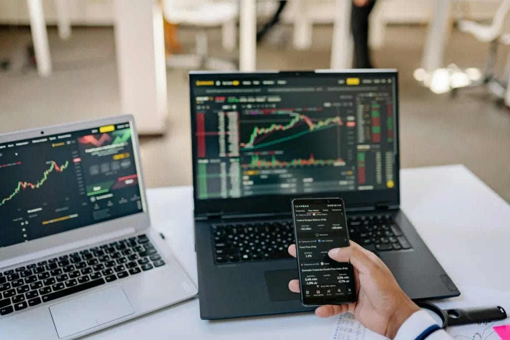 Person analyzing financial charts on a smartphone and two laptops showing trading graphs and market data.