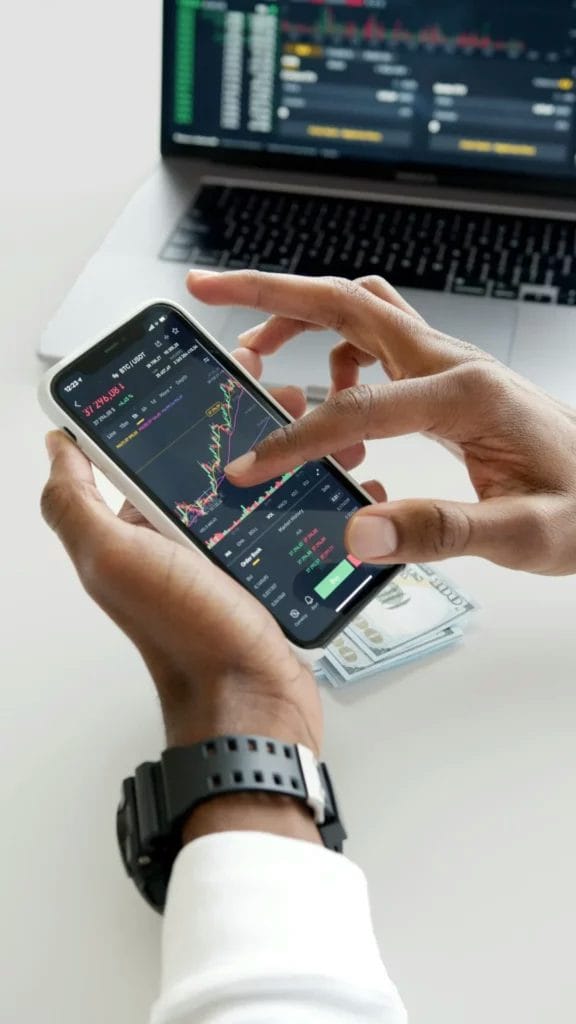 A person holds a smartphone with a trading app, a laptop with financial data in the background, and dollar bills on the desk.
