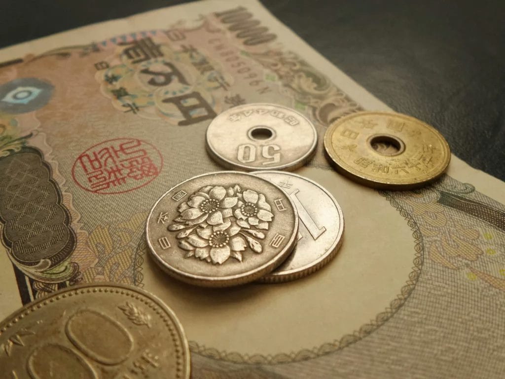 The image shows Japanese yen currency, including a 10,000 yen banknote and various coins (50 yen, 10 yen, 5 yen, and 1 yen). The banknote features intricate designs and Japanese characters, while the coins display floral patterns and denominations.